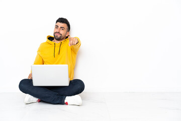 Caucasian man sitting on the floor with his laptop points finger at you with a confident expression