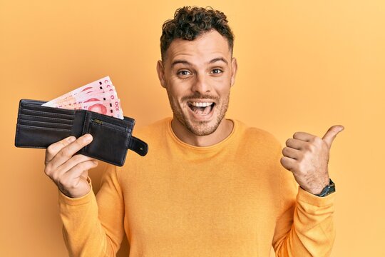 Young hispanic man holding wallet with yuan chinese banknotes pointing thumb up to the side smiling happy with open mouth
