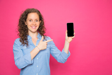 portrait color young redhead woman smiling pointing mobile phone with denim shirt on pink background looking at camera