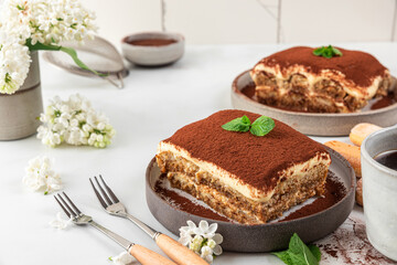 Homemade traditional italian dessert tiramisu in a plate with coffee cup, dessert forks and flowers on white background