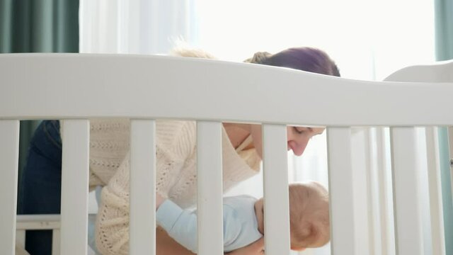 Caring young mother holding her baby and putting in cradle. Concept of parenting, family happiness and baby development.