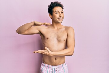 Young handsome man wearing swimwear shirtless gesturing with hands showing big and large size sign, measure symbol. smiling looking at the camera. measuring concept.