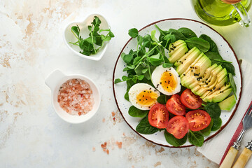 Avocado, cherry tomato, spinach and chicken egg, microgreens peas and black sesame seeds fresh salad in bowl on white stone table background. Healthy breakfast food concept. Top view.
