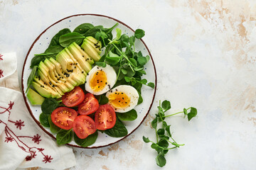 Avocado, cherry tomato, spinach and chicken egg, microgreens peas and black sesame seeds fresh salad in bowl on white stone table background. Healthy breakfast food concept. Top view.