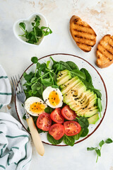 Avocado, cherry tomato, spinach and chicken egg, microgreens peas and black sesame seeds fresh salad in bowl on white stone table background. Healthy breakfast food concept. Top view.