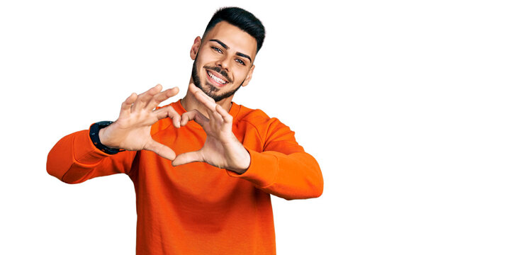 Young hispanic man with beard wearing casual orange sweater smiling in love doing heart symbol shape with hands. romantic concept.