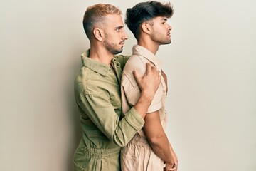 Homosexual gay couple standing together wearing casual jumpsuit looking to side, relax profile pose with natural face and confident smile.
