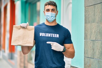 Young hispanic volunteer man wearing medical mask pointing with finger to delivery bag at the city.