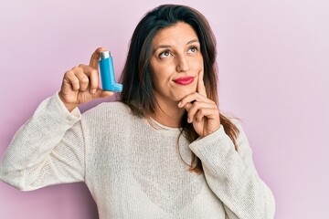 Beautiful hispanic woman holding medical asthma inhaler serious face thinking about question with hand on chin, thoughtful about confusing idea - obrazy, fototapety, plakaty