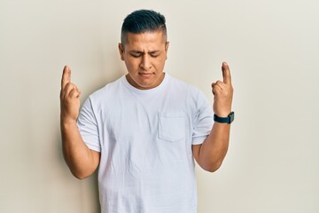 Fototapeta premium Young latin man wearing casual white t shirt gesturing finger crossed smiling with hope and eyes closed. luck and superstitious concept.