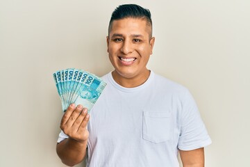 Young latin man holding 100 brazilian real banknotes looking positive and happy standing and...