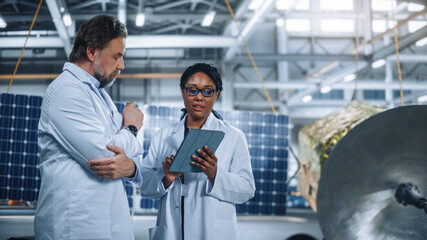 Two Engineers Talk, Use Tablet Computer while Working on Satellite Construction. Aerospace Agency Manufacturing Facility: Scientists Build, Assemble Spacecraft for Space Exploration Mission