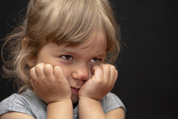 Portrait of a sad little girl with a scratched nose on a dark background, holding her face in her hands.