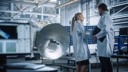 Engineer and Technician Talk, Use Tablet Computer while Working on Satellite Construction. Aerospace Agency Manufacturing Facility: Scientists Build, Assemble Spacecraft for Space Exploration Mission