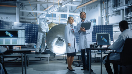 Happy Engineer and Technician Talk, Use Laptop Computer while Working on Satellite Construction. Aerospace Agency Manufacturing Facility: Scientists Assemble Spacecraft for Space Exploration Mission