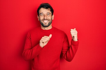 Handsome man with beard wearing casual red sweater pointing to the back behind with hand and thumbs...