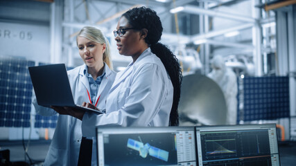 Two Female Engineers Talk, Use Computer working at Aerospace Satellite Manufacturing Facility....
