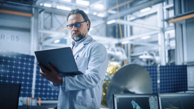Male Engineer Confident and Focused Thinking, working on Laptop at Aerospace Satellite Manufacturing Facility. Top World Scientists Doing Science and Technology Research in Space Program
