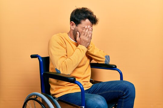 Handsome man with beard sitting on wheelchair with sad expression covering face with hands while crying. depression concept.