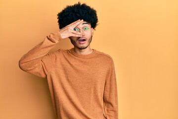 Fototapeta na wymiar Young african american man with afro hair wearing casual winter sweater peeking in shock covering face and eyes with hand, looking through fingers with embarrassed expression.