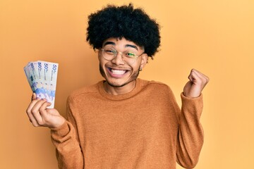 Young african american man with afro hair holding 50 mexican pesos banknotes screaming proud, celebrating victory and success very excited with raised arm