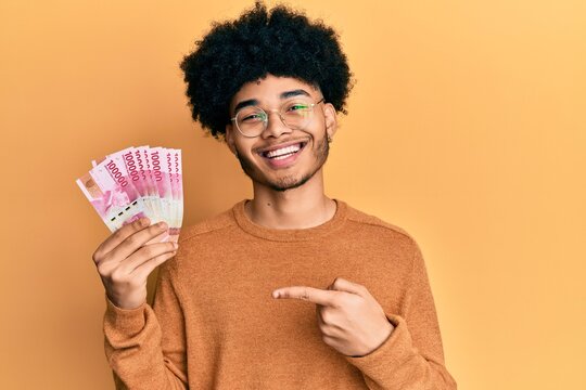 Young african american man with afro hair holding 100000 indonesian rupiah smiling happy pointing with hand and finger