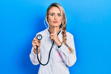 Beautiful young blonde doctor woman holding stethoscope thinking concentrated about doubt with finger on chin and looking up wondering