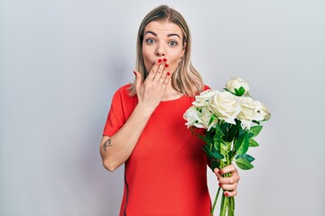 Beautiful caucasian woman holding bouquet of white flowers covering mouth with hand, shocked and...
