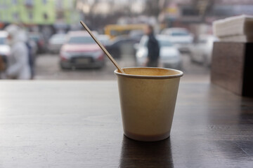 used disposable paper cup with a wooden stick stands on the windowsill against the background of a transparent window.