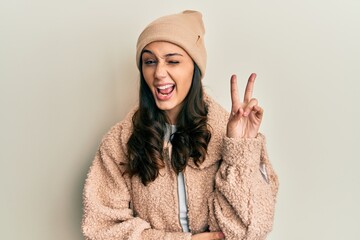 Young hispanic woman wearing wool sweater and winter hat smiling with happy face winking at the...