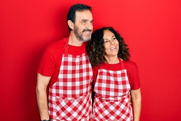 Middle age couple of hispanic woman and man wearing cook apron looking away to side with smile on face, natural expression. laughing confident.