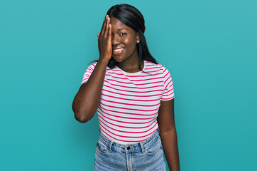 African young woman wearing casual striped t shirt covering one eye with hand, confident smile on face and surprise emotion.