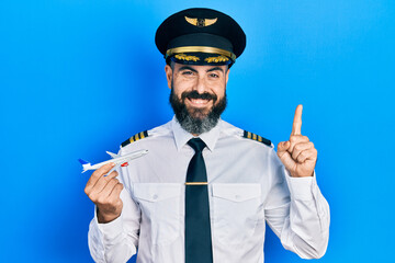 Young hispanic man wearing pilot uniform holding plane toy smiling with an idea or question pointing finger with happy face, number one - obrazy, fototapety, plakaty