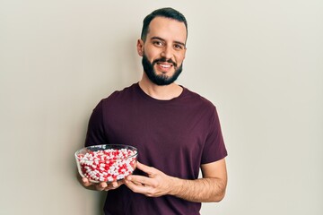 Young man with beard holding bowl full of pills looking positive and happy standing and smiling with a confident smile showing teeth