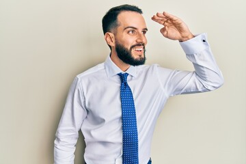 Young man with beard wearing business tie very happy and smiling looking far away with hand over head. searching concept.