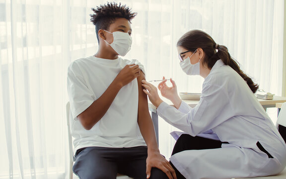 African Teen Man Wearing Face Mask, Getting Vaccination To Protect Or Prevent Virus, Getting Scary Vaccine Injection While Female Caucasian Doctor Preparing Syringe To Vaccinate Patient At Hospital.