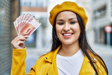 Young latin girl smiling happy holding norway krone banknotes at the city.