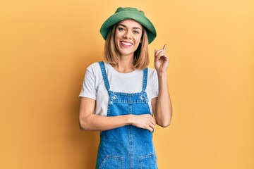 Young caucasian blonde woman wearing denim jumpsuit and hat with 90s style with a big smile on...
