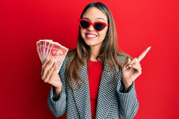 Obraz premium Young brunette woman holding 20 israel shekels banknotes smiling happy pointing with hand and finger to the side