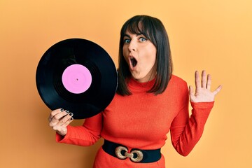 Young hispanic woman holding vinyl disc scared and amazed with open mouth for surprise, disbelief face