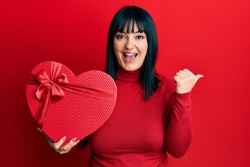 Young hispanic woman holding valentine gift pointing thumb up to the side smiling happy with open mouth