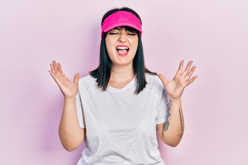 Young hispanic woman wearing sportswear and sun visor cap celebrating mad and crazy for success with arms raised and closed eyes screaming excited. winner concept