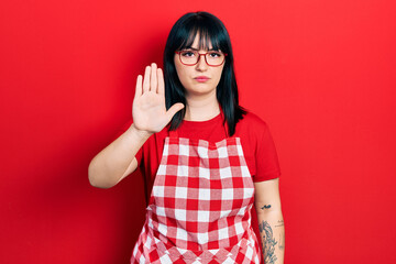 Young hispanic woman wearing cook apron and glasses doing stop sing with palm of the hand. warning expression with negative and serious gesture on the face.