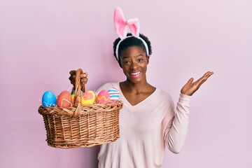 Young african american girl wearing cute easter bunny ears holding basket with painted eggs celebrating victory with happy smile and winner expression with raised hands