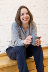 Vertical portrait of young brunette woman sitting on wooden flooring with cup of coffee in her hand on background of white brick wall. The woman smiles and holds a ceramic gray mug in her hand.