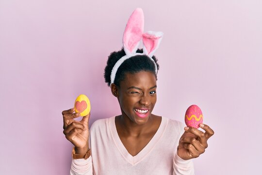 Young african american girl wearing cute easter bunny ears holding painted eggs winking looking at the camera with sexy expression, cheerful and happy face.