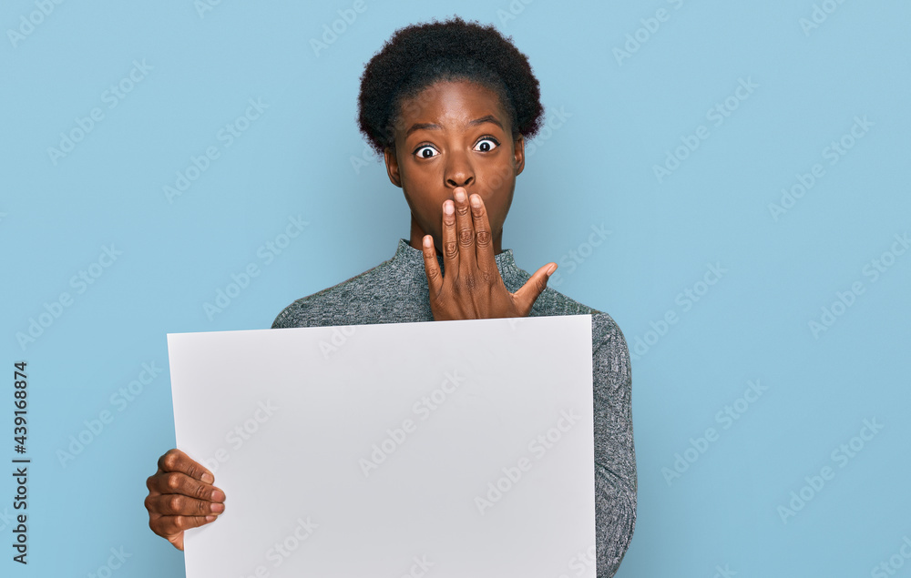 Poster Young african american girl holding blank empty banner covering mouth with hand, shocked and afraid for mistake. surprised expression