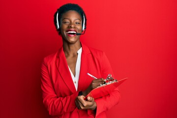 Young african american girl wearing call center agent headset writing on clipboard smiling and...