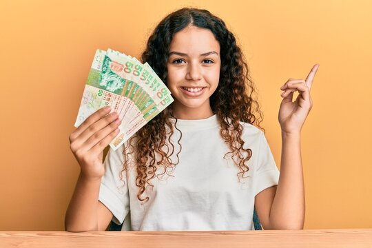 Teenager hispanic girl holding 50 hong kong dollars banknotes smiling happy pointing with hand and finger to the side