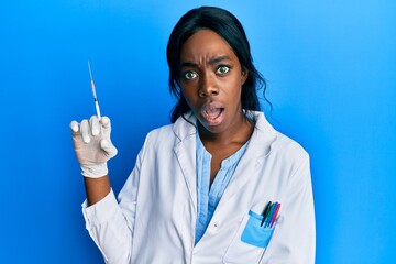 Young african american woman wearing scientist uniform holding syringe scared and amazed with open mouth for surprise, disbelief face